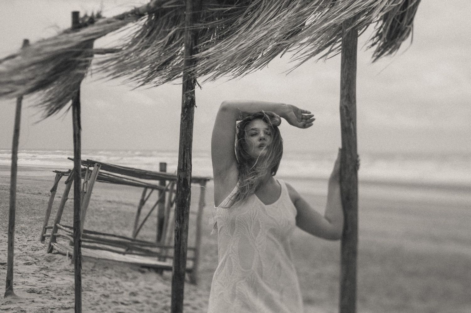 A vintage black and white photo of someone under a thatched beach shelter.