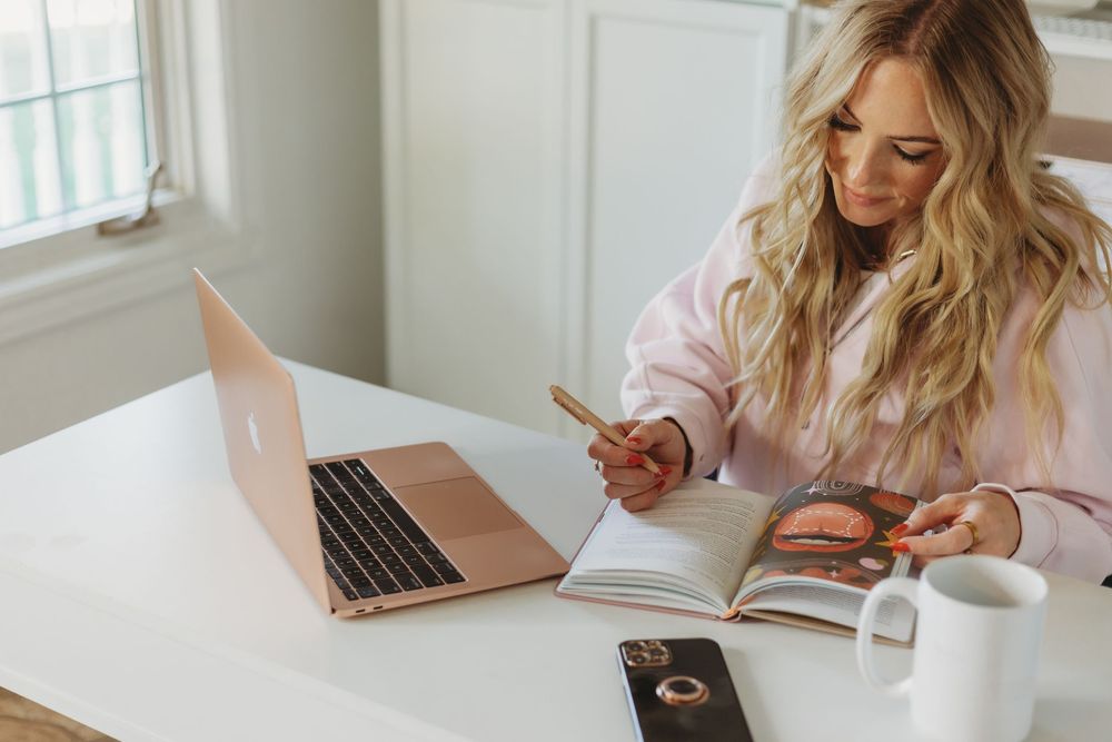 Someone in a pink sweater works at a bright white desk with a laptop and notebook.