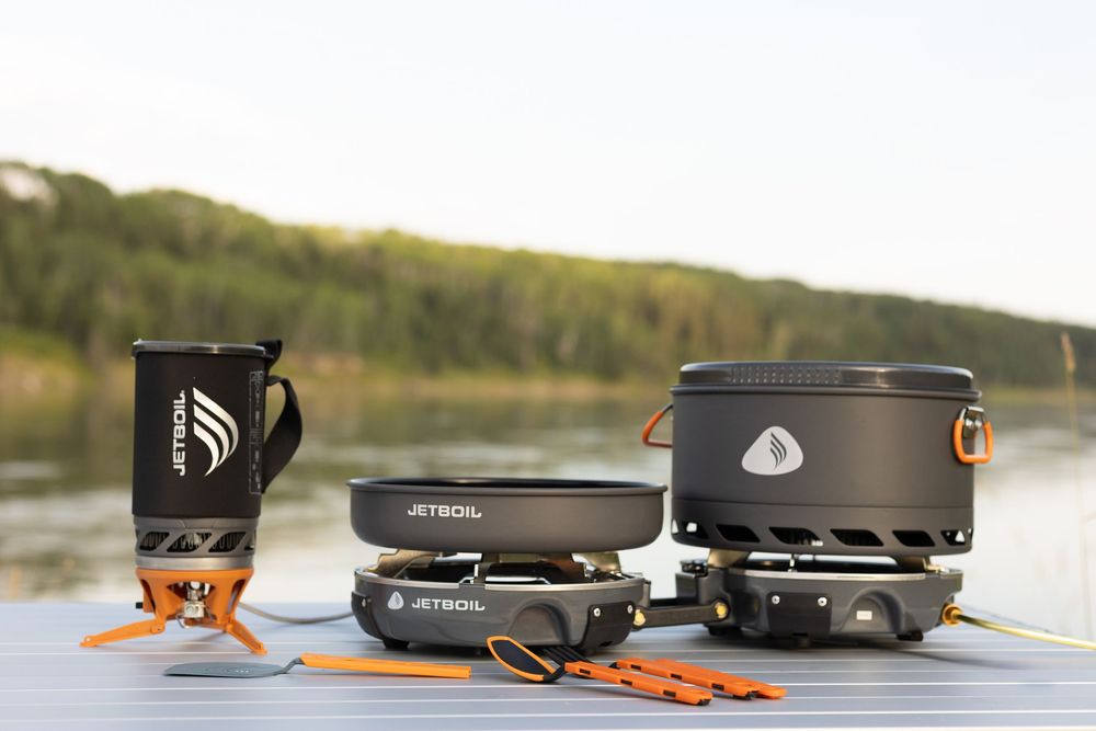 A series of portable camping stoves and cooking equipment arranged on a wooden surface outdoors.