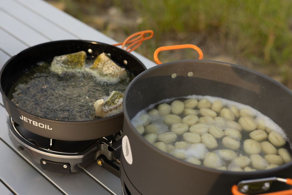 Two large outdoor cooking pans on a table, one frying food and another filled with boiling potatoes.