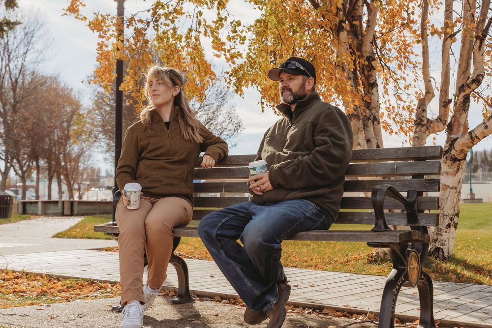 Two people sit on a park bench surrounded by autumn trees with fallen leaves on a crisp fall day.
