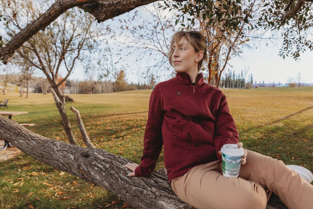 A person in a burgundy sweater sits on a fallen tree trunk in an autumn field with a coffee cup.
