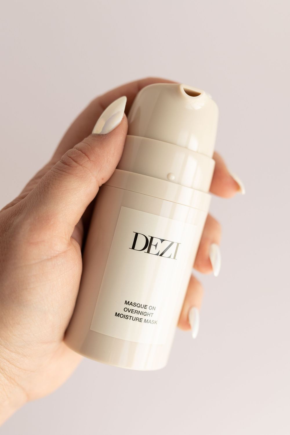 Hand holding a minimalist white skincare product container with 'DEZ' branding against a light background.