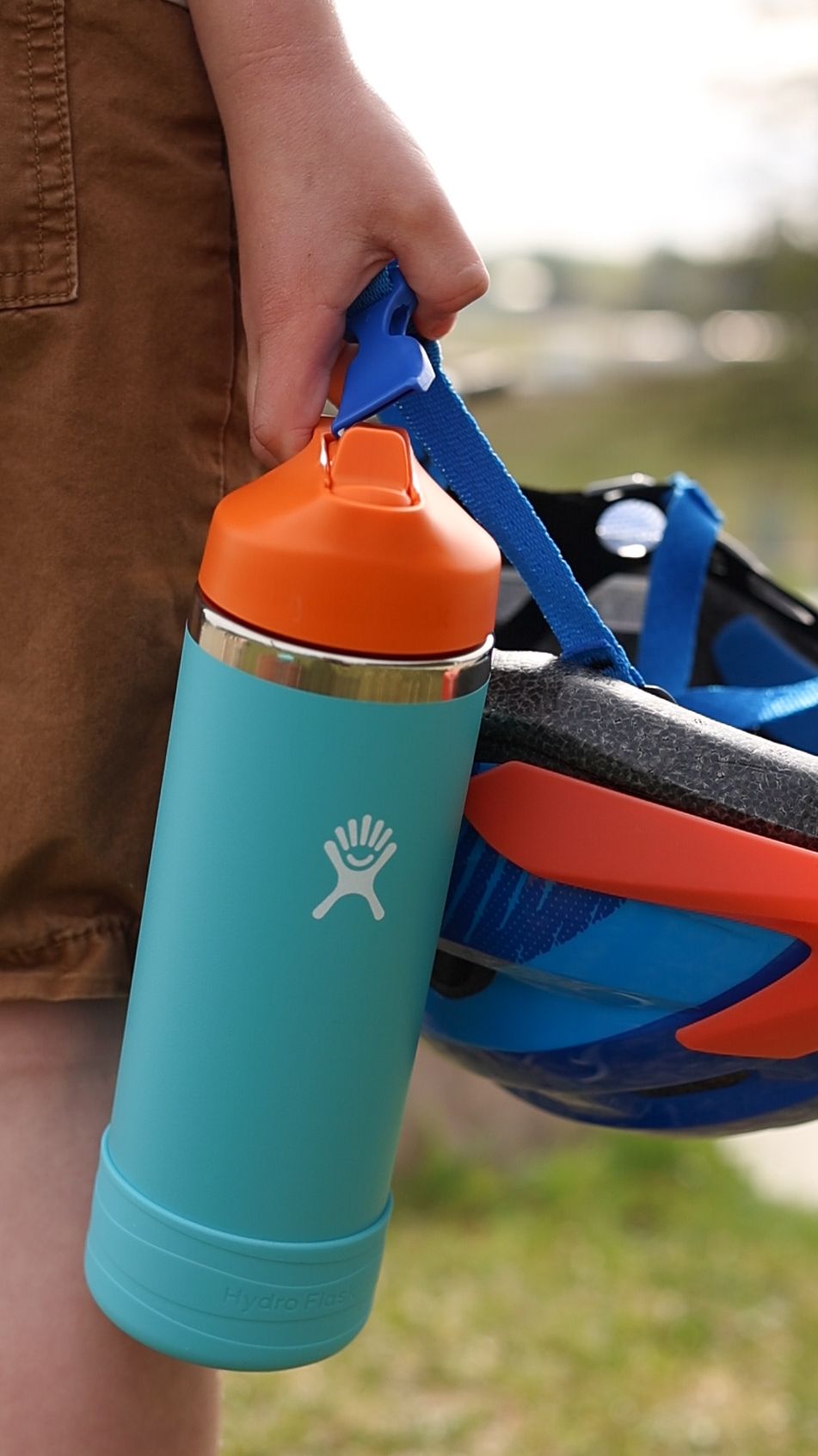 A hand holds a teal Hydro Flask water bottle with an orange lid next to a blue and orange bicycle helmet.