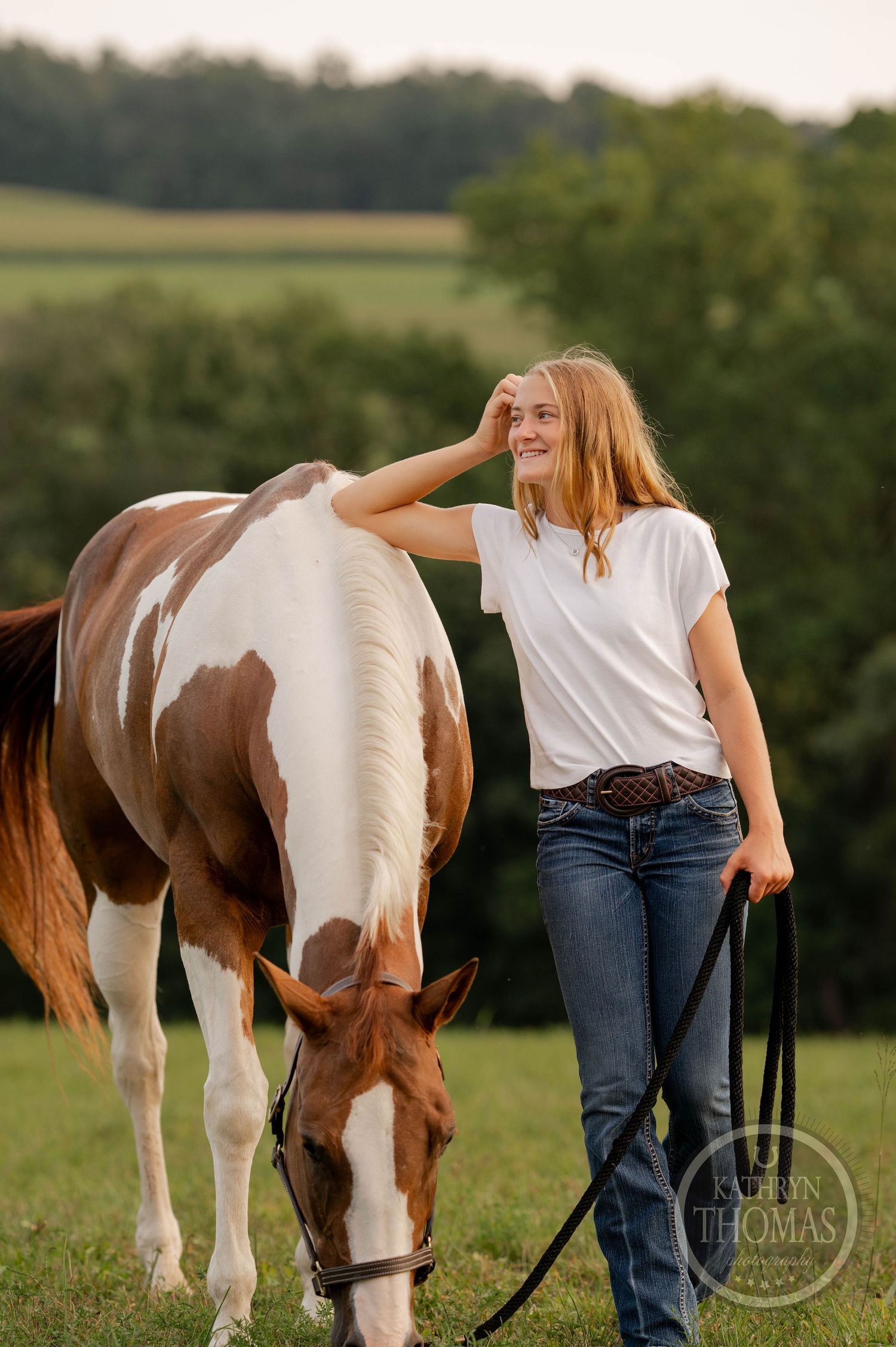 A paint horse grazes peacefully in a golden-lit field while someone stands beside it.