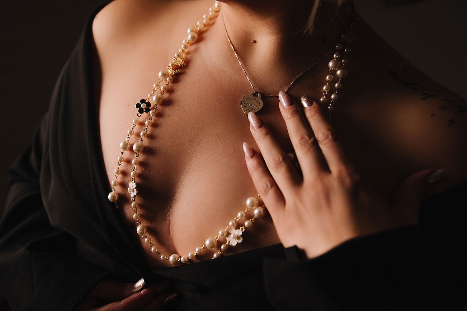 Elegant pearl necklace and well-manicured hands against black fabric in moody lighting create a luxurious fashion detail shot.