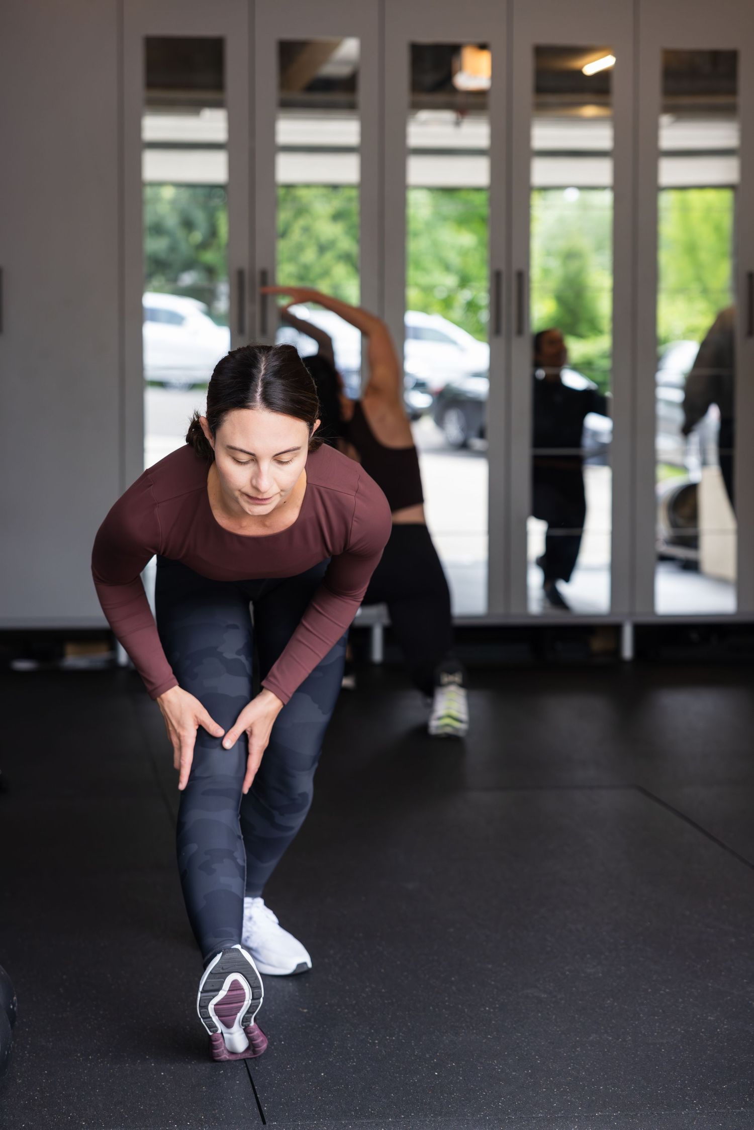 Person in a burgundy long sleeve top and dark pants stretches on a gym floor with windows visible in background.