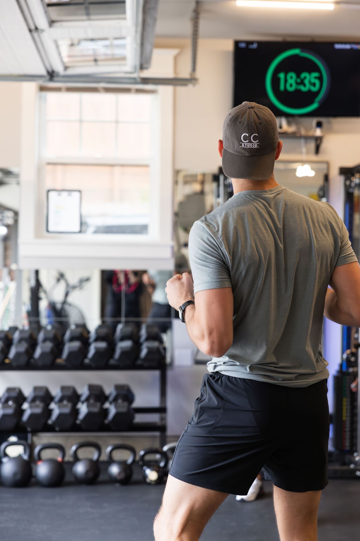 Person in gray t-shirt and black shorts working out in a gym with kettlebells and a digital clock display.