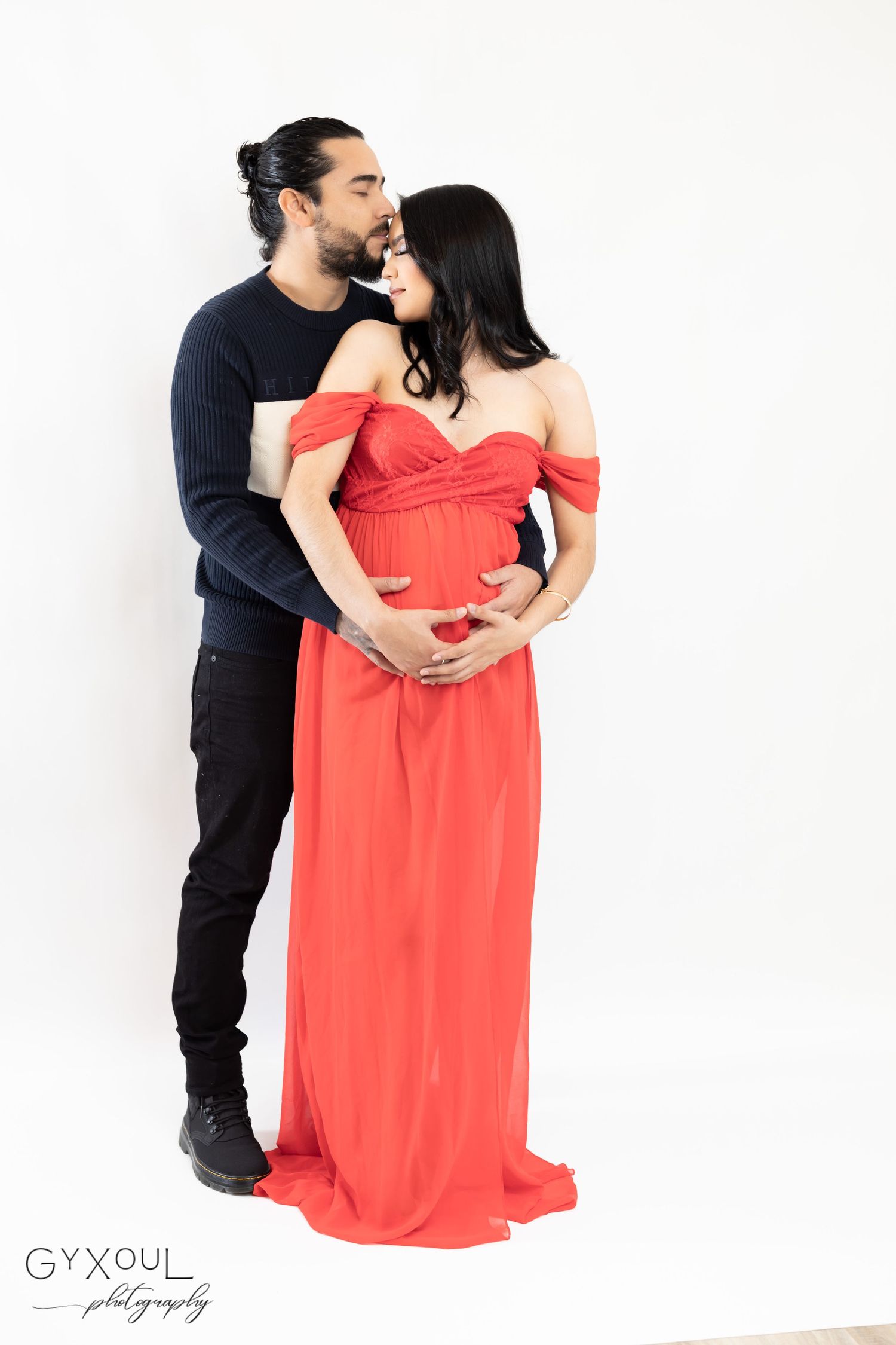 Elegant maternity portrait with couple embracing against white backdrop.