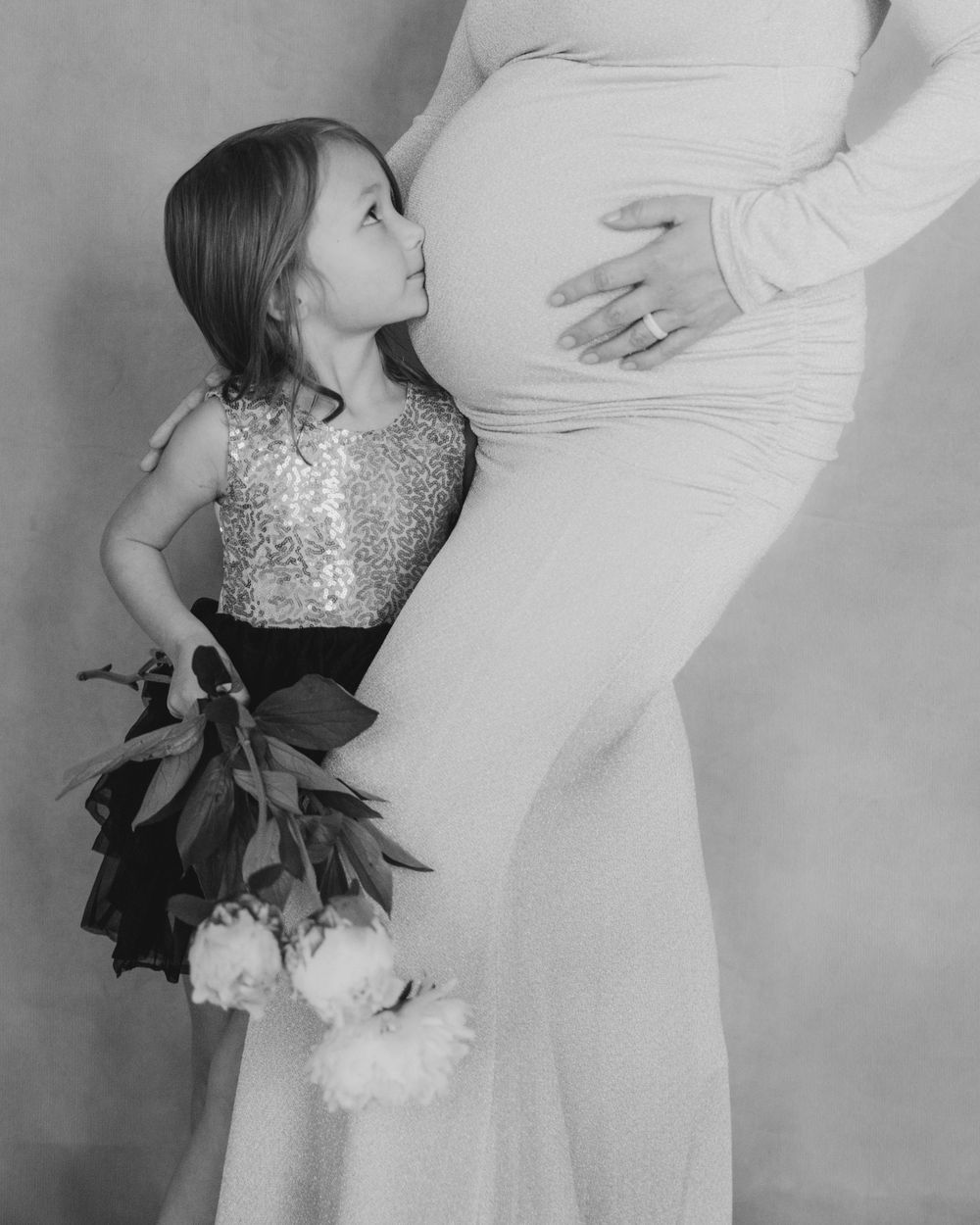 A touching black and white portrait of a young girl in a sparkly dress looking at a pregnant womans belly while holding flowers.