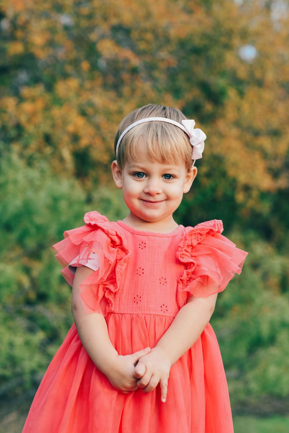 A young child wearing a coral ruffle dress and white floral headband poses outdoors against an autumn backdrop.