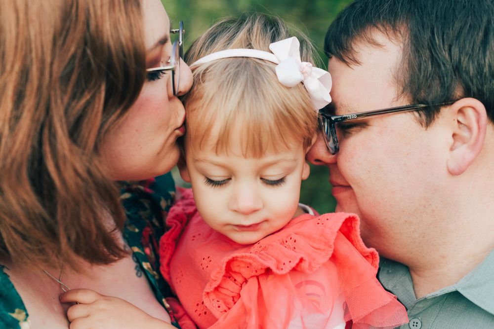 Young child in a coral dress smiling while being embraced by parents wearing glasses outdoors.
