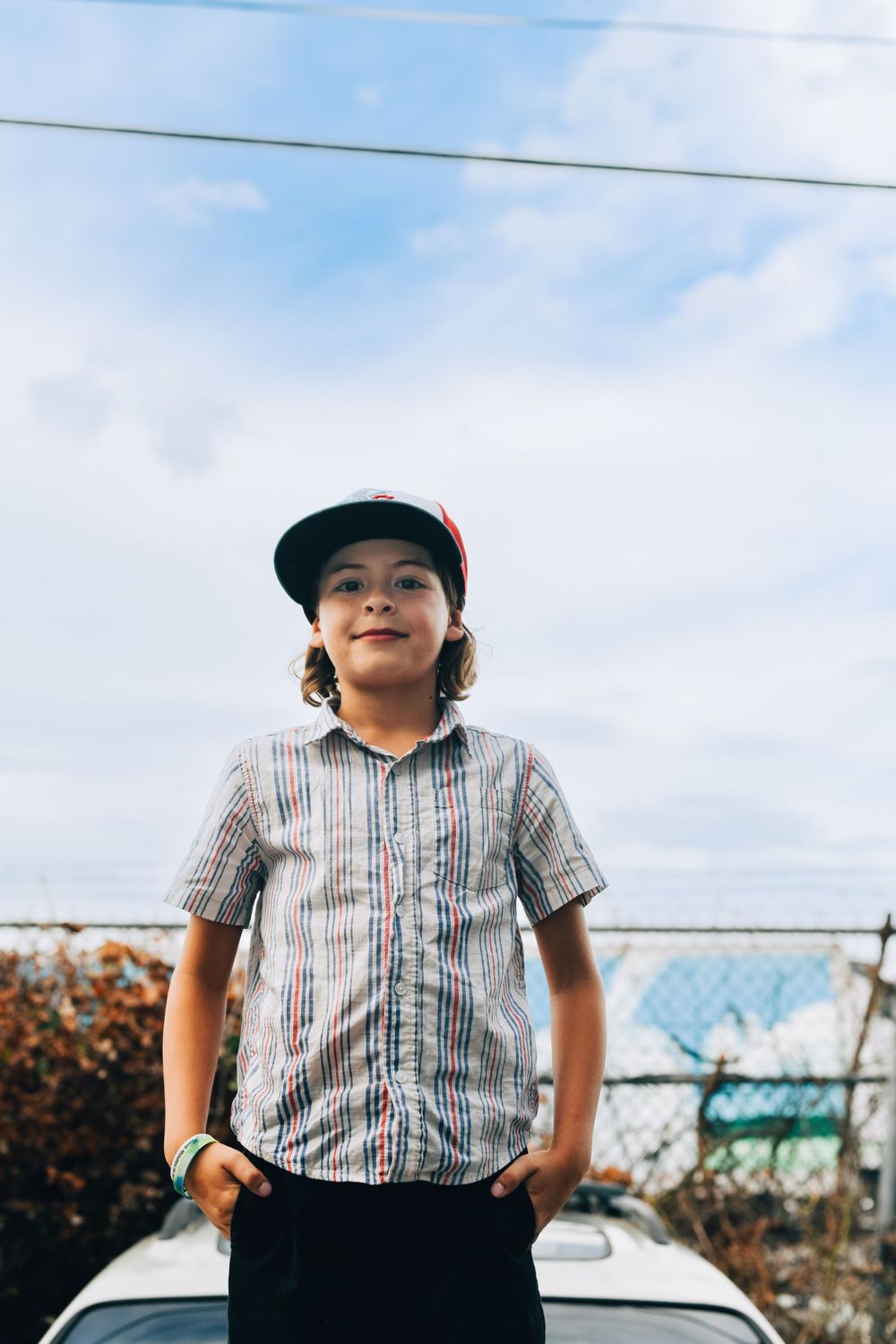 Young person wearing baseball cap and striped shirt poses outdoors in front of parked car under cloudy sky.