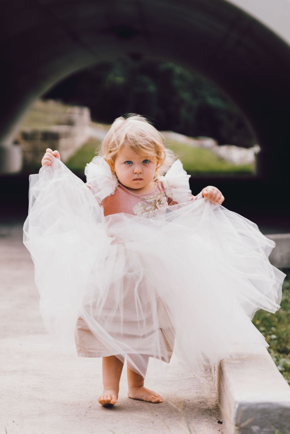 Small child wearing an elegant white tulle dress holding up the skirt while standing on a pathway.