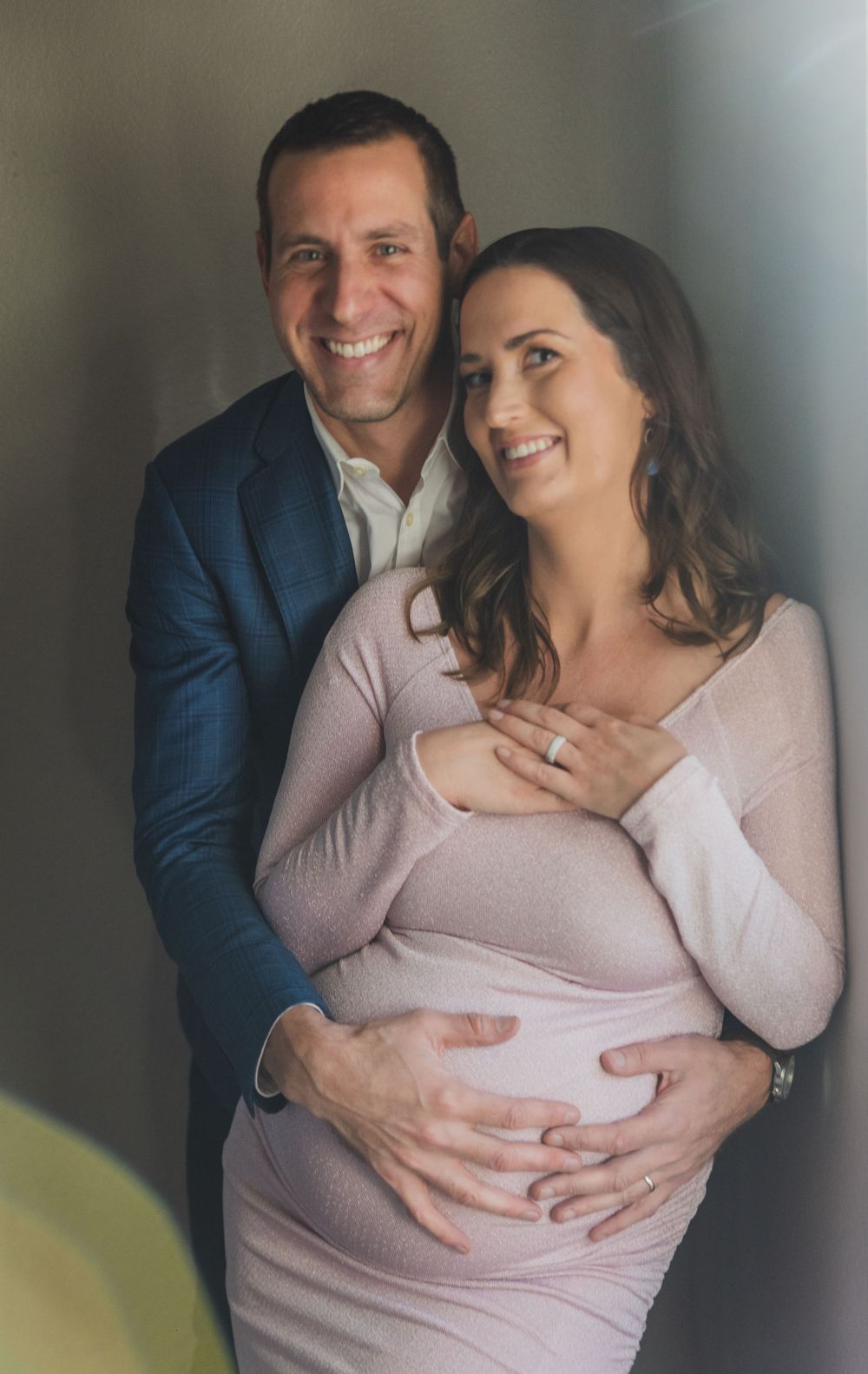 A couple in pink and blue formal wear embraces during a maternity photo shoot, with hands resting on her pregnant belly.