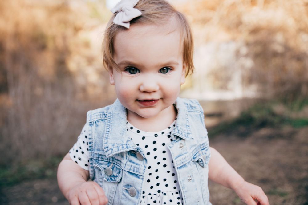 Toddler wearing denim vest and polka dot dress smiles outdoors in warm natural lighting.