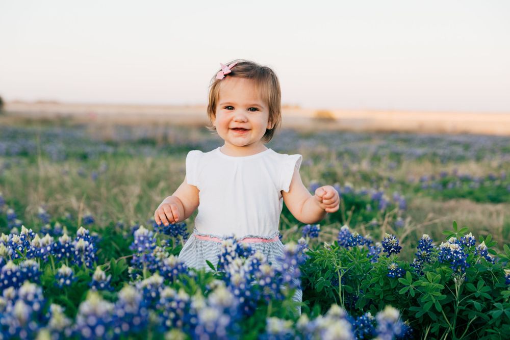 Small child in white dress sits among blooming bluebonnets in a Texas field during golden hour.