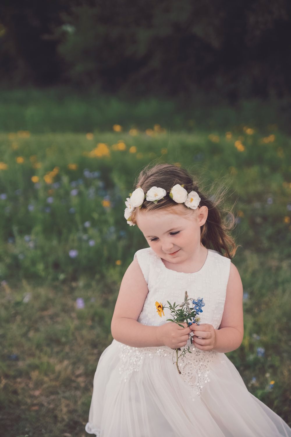 Child in white dress and floral crown holds small bouquet of wildflowers in sunny meadow.