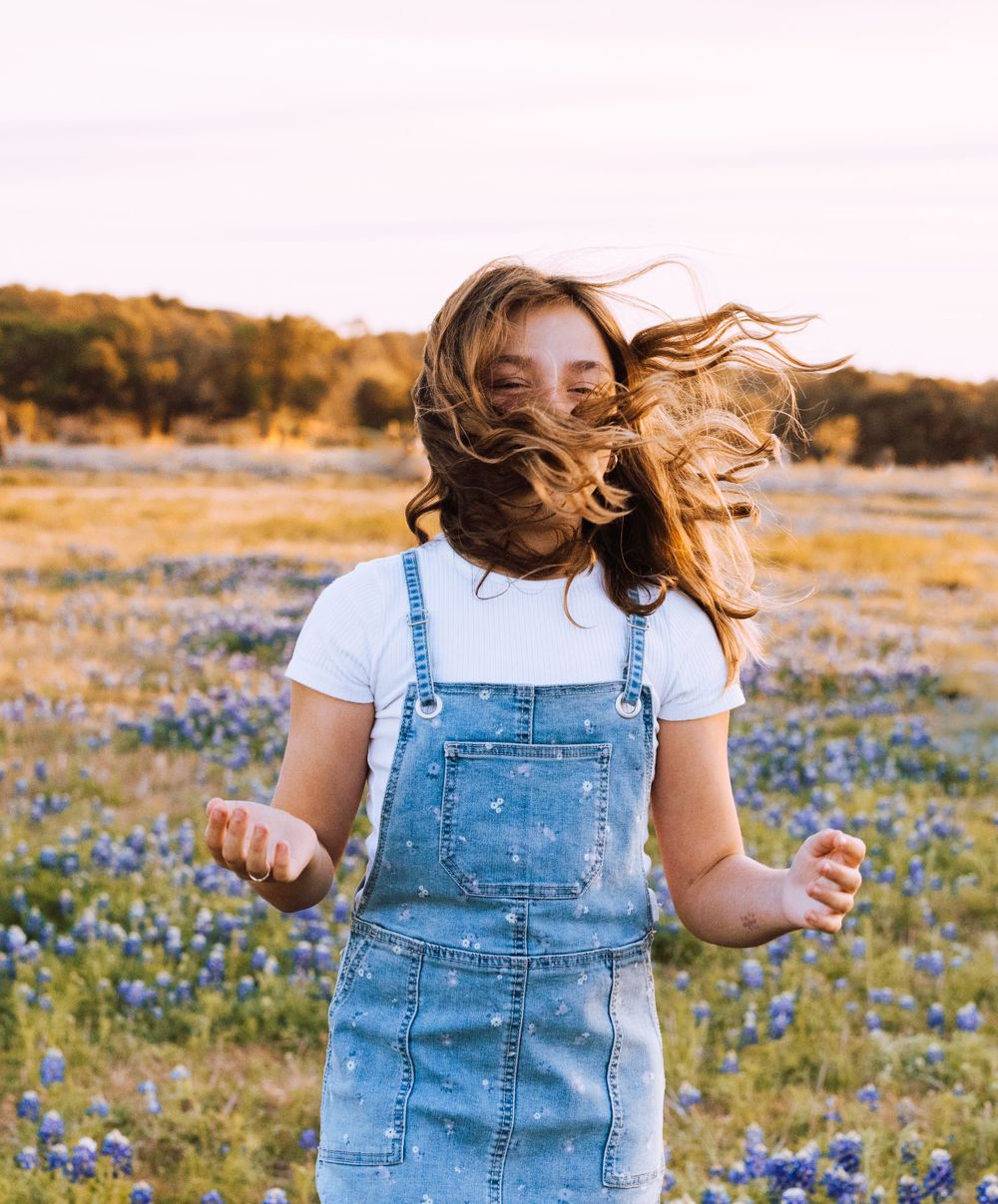Person with flowing hair dances in a field of blue wildflowers at golden hour wearing denim overalls and white shirt.