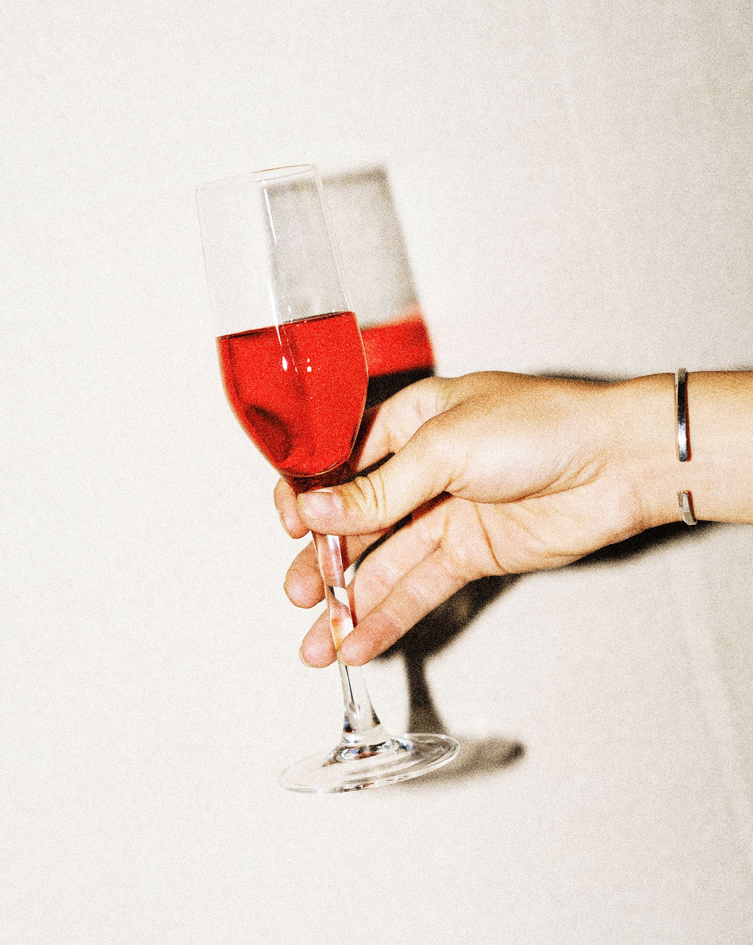 A hand wearing delicate bracelets holds a wine glass filled with red wine against a white background.