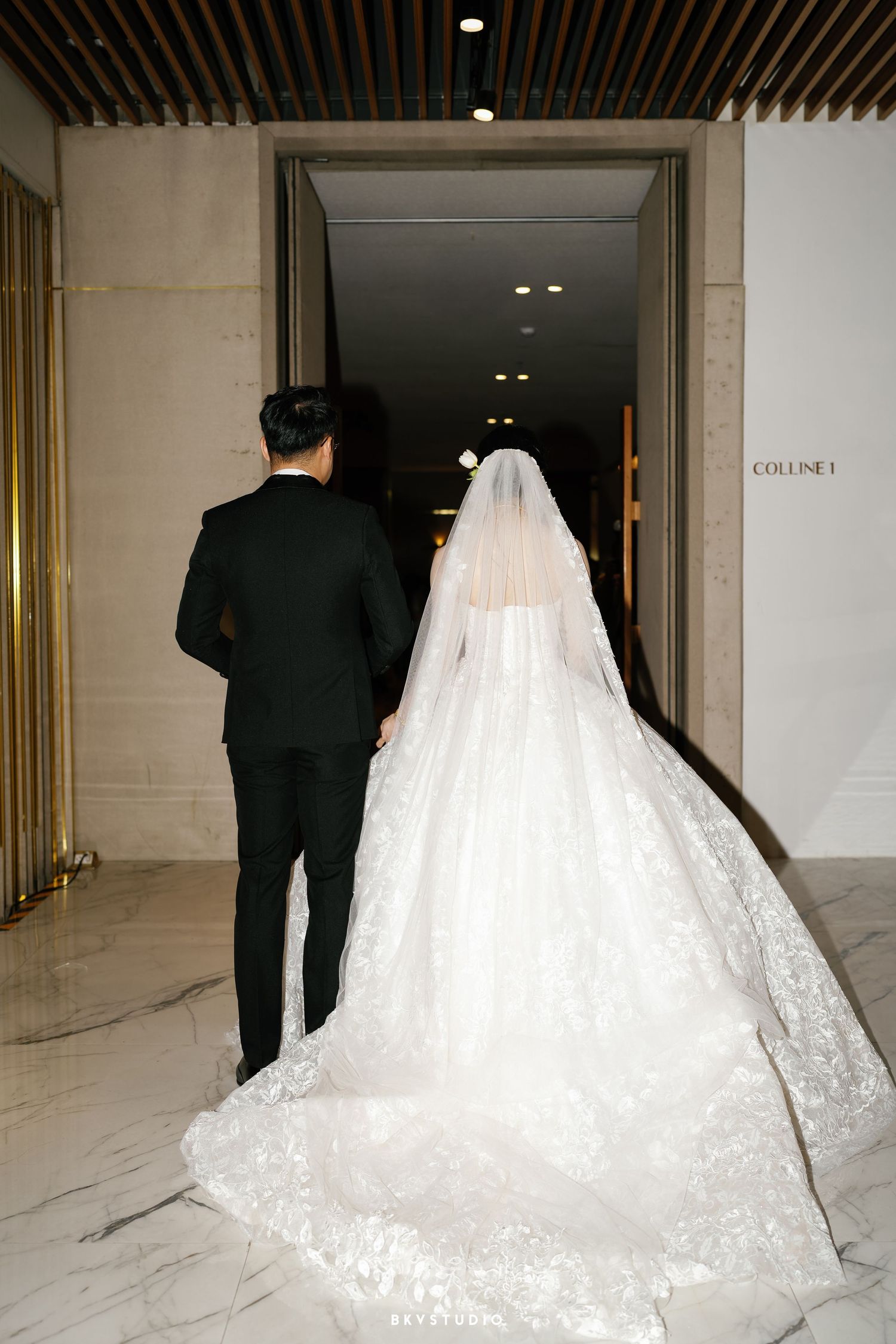 A bride in a flowing lace wedding gown with long train walks down a marble hallway with someone in a black suit.