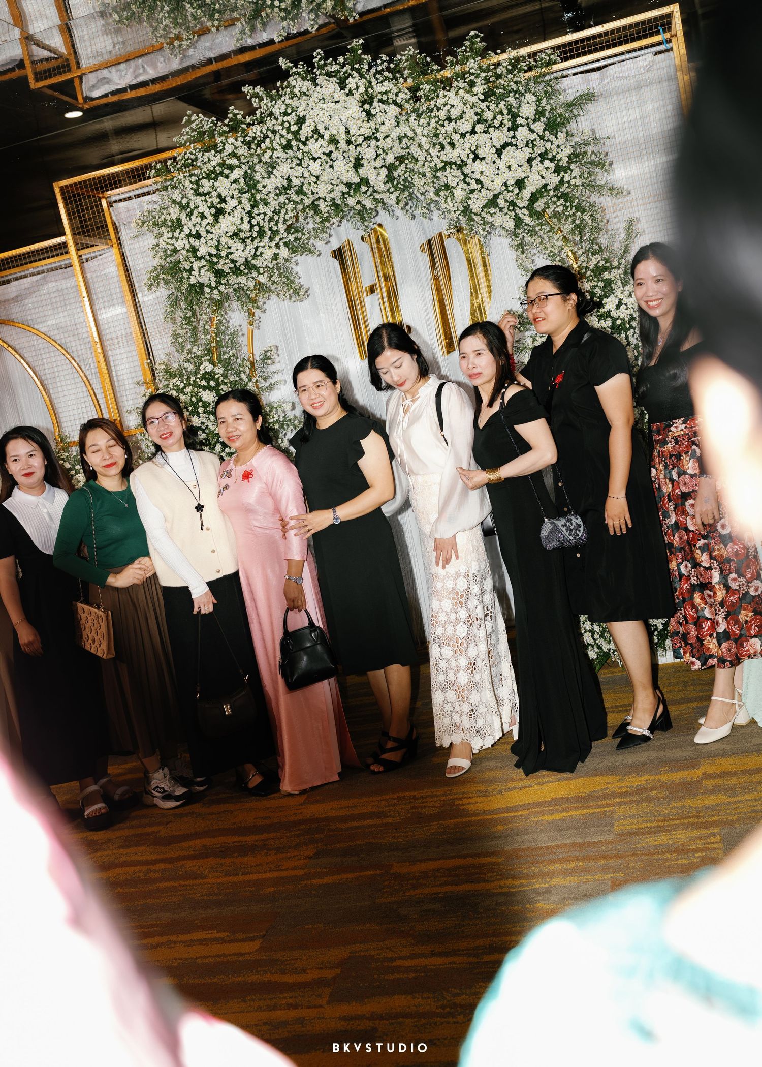 A group of women in formal attire stand together at an event under white floral decorations and gold accents.