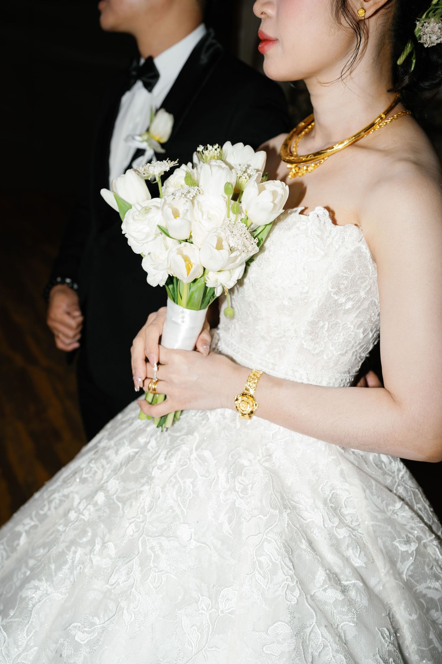 Wedding couple with white bridal bouquet and elegant gold jewelry details against dark background.