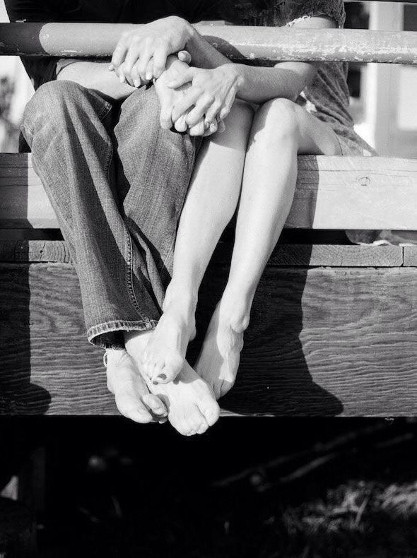 A black and white photo showing two pairs of bare feet intimately intertwined on a wooden bench or dock.