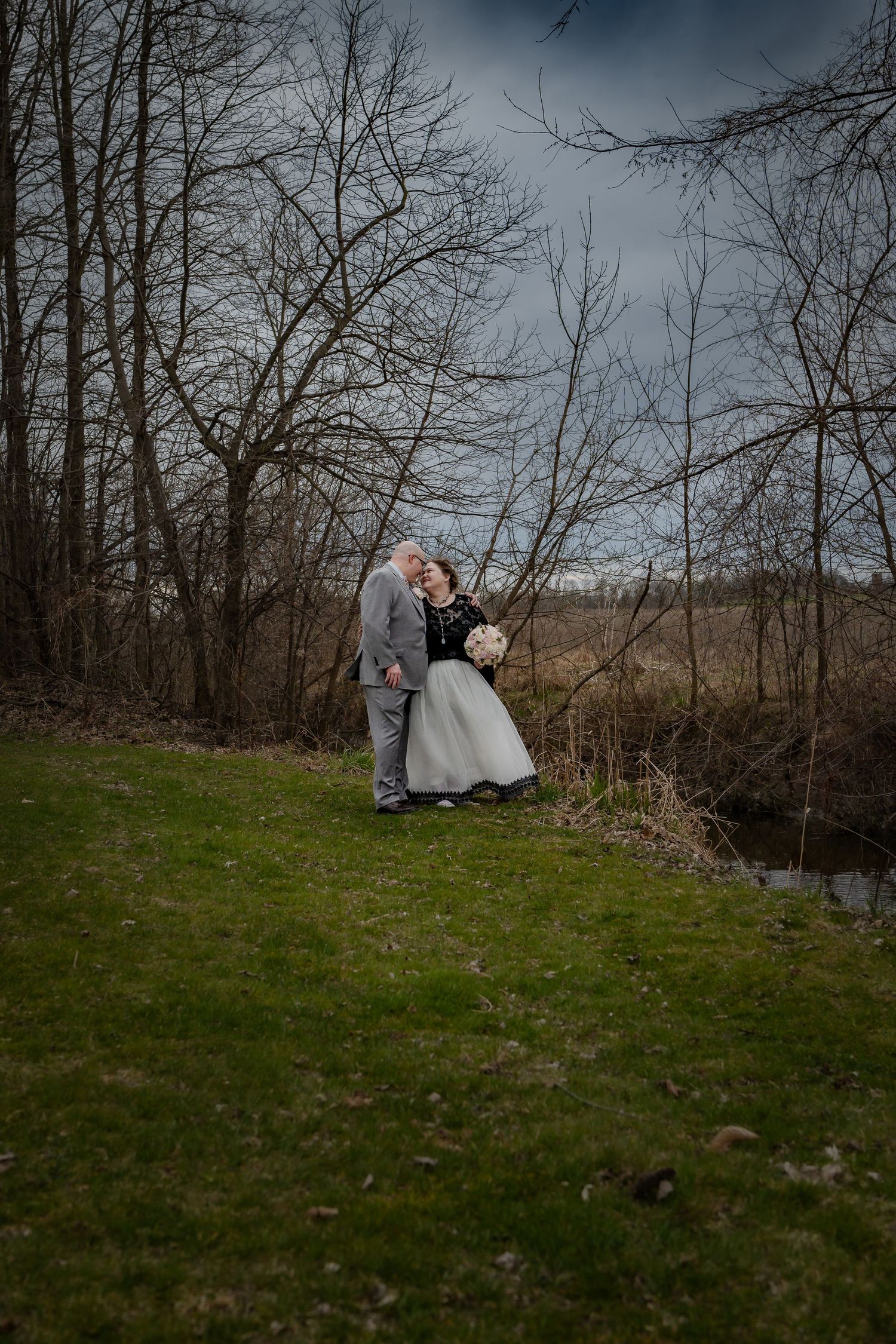 A couple in wedding attire embraces on a grassy hillside near a stream with bare winter trees in the background.