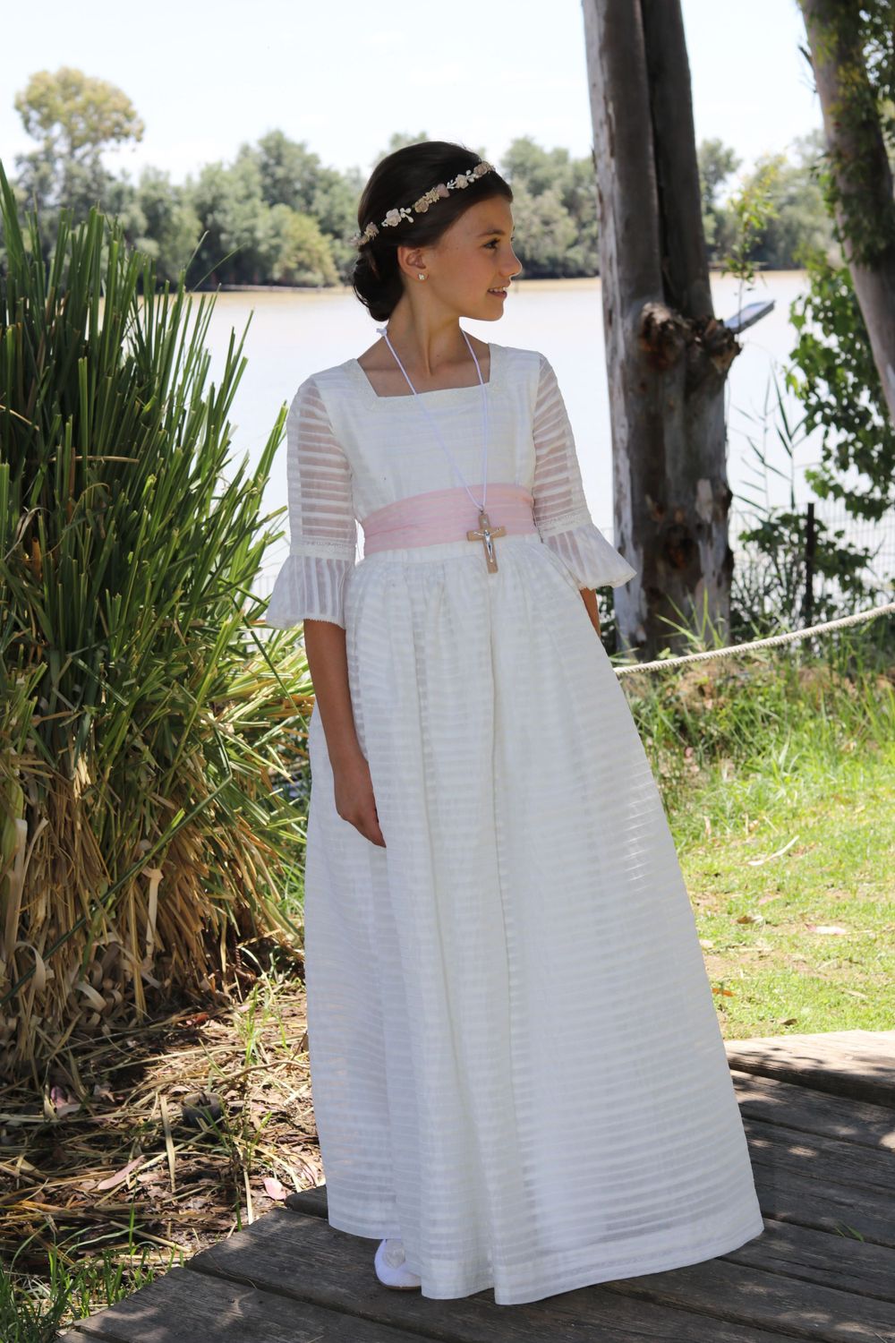 A white floor-length dress with striped texture and pink sash stands elegantly by a lake surrounded by reeds and trees.