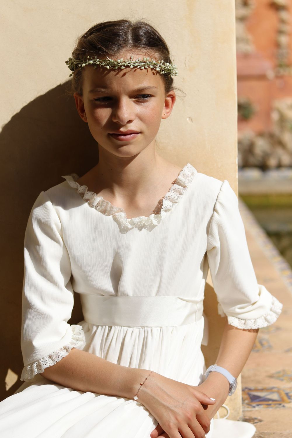 A young person in a traditional white communion dress with delicate lace trim and a floral crown sitting in sunlight.