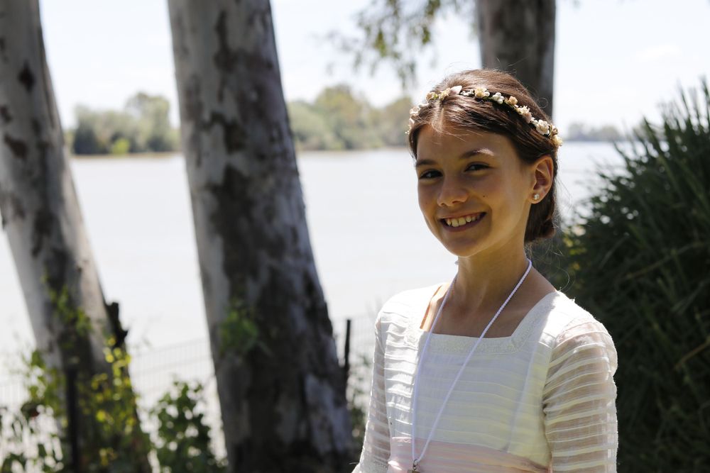 Someone in a white dress and floral headpiece smiles while standing by a lake with tree trunks in the background.