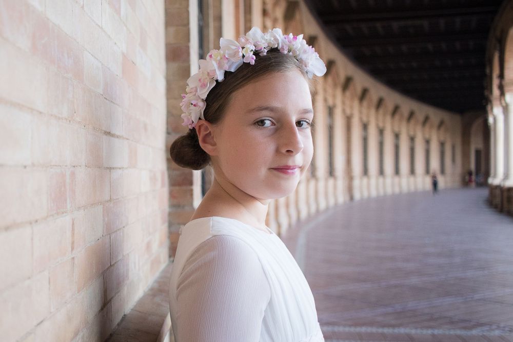 A young figure wearing white stands in an ornate stone corridor while wearing a delicate floral crown.