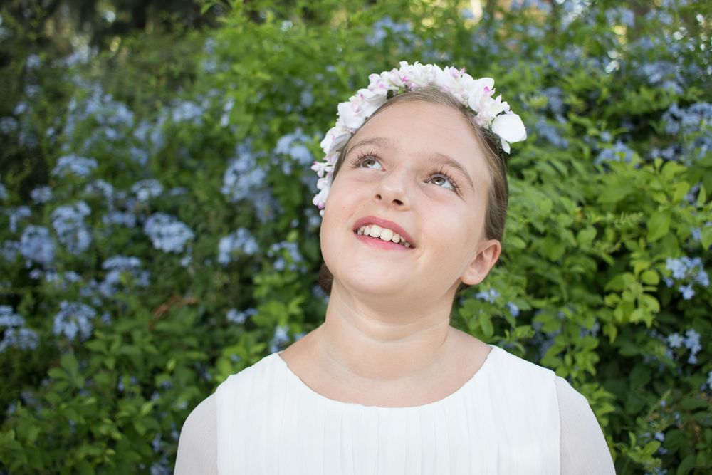 A white-robed figure with a flower crown smiles upward against a backdrop of blue flowers and green foliage.