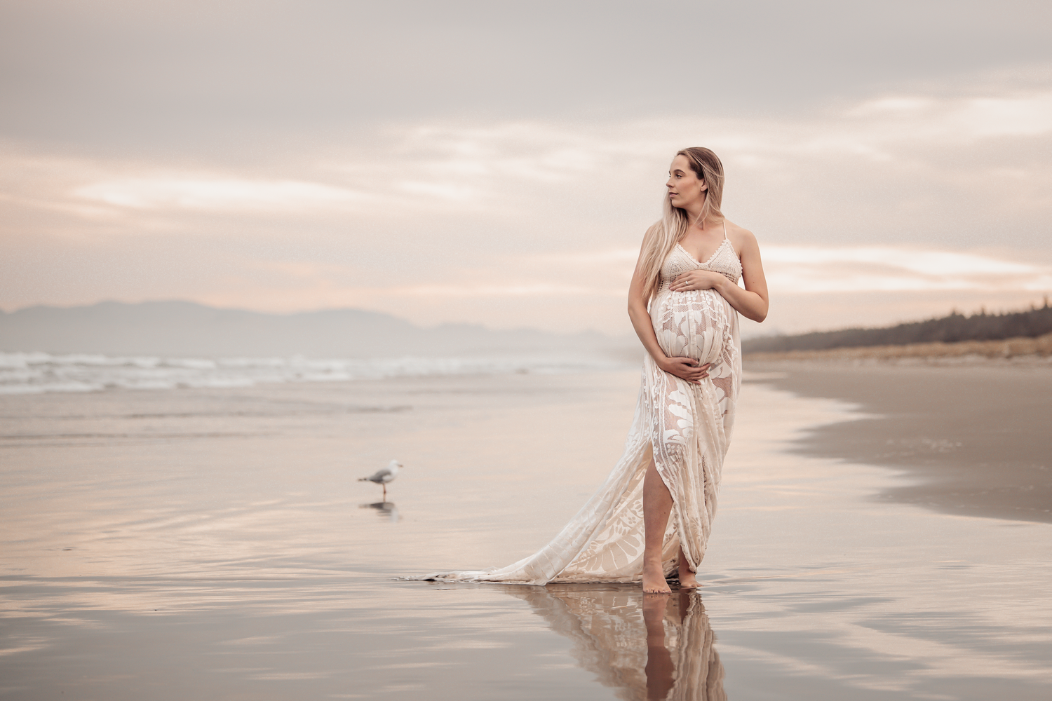 Maternity portrait of woman in white dress standing in shallow ocean waves at sunset.