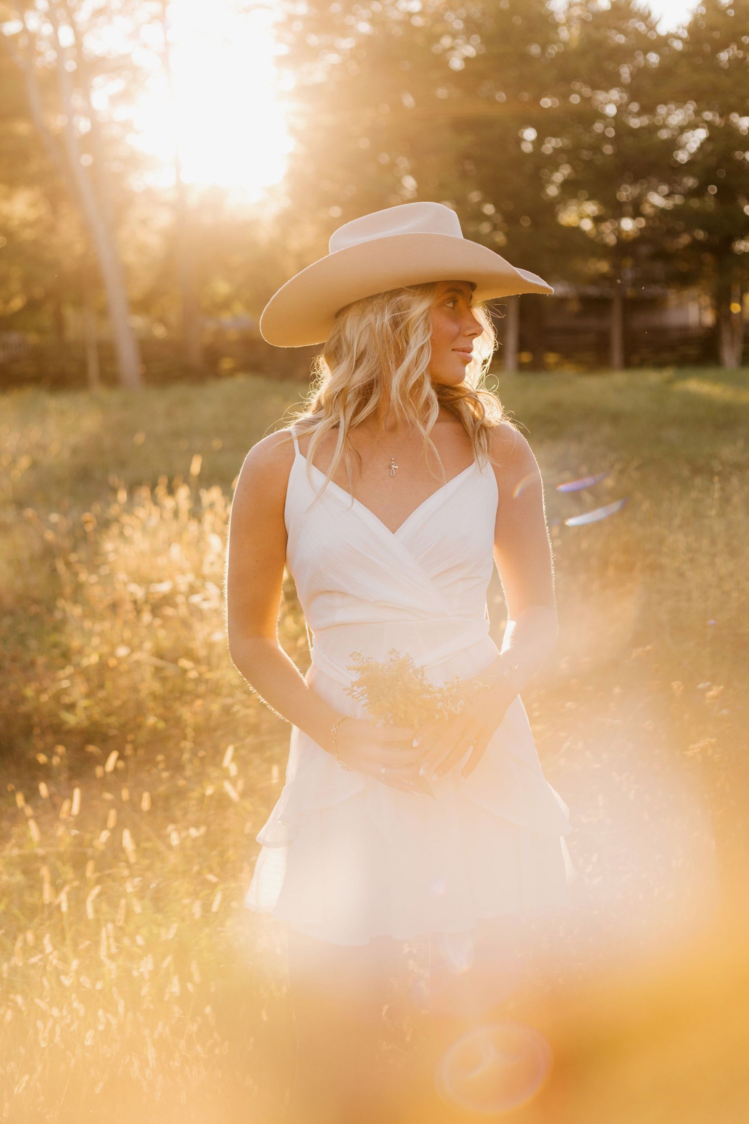 A person in a white dress and cowboy hat stands in a golden wheat field at sunset with dramatic lens flare.