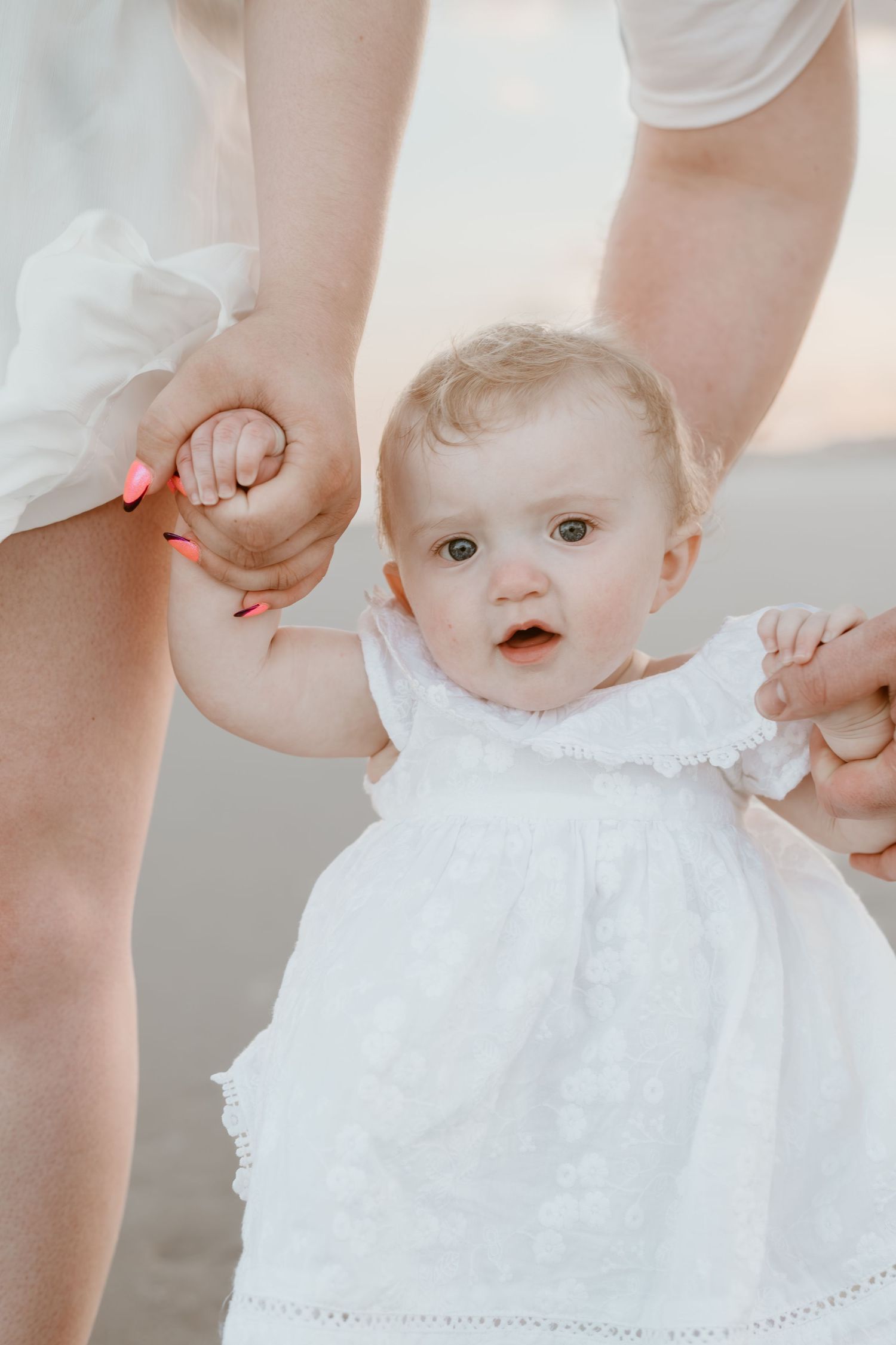 Baby in white lace dress being supported while learning to take first steps on the beach.
