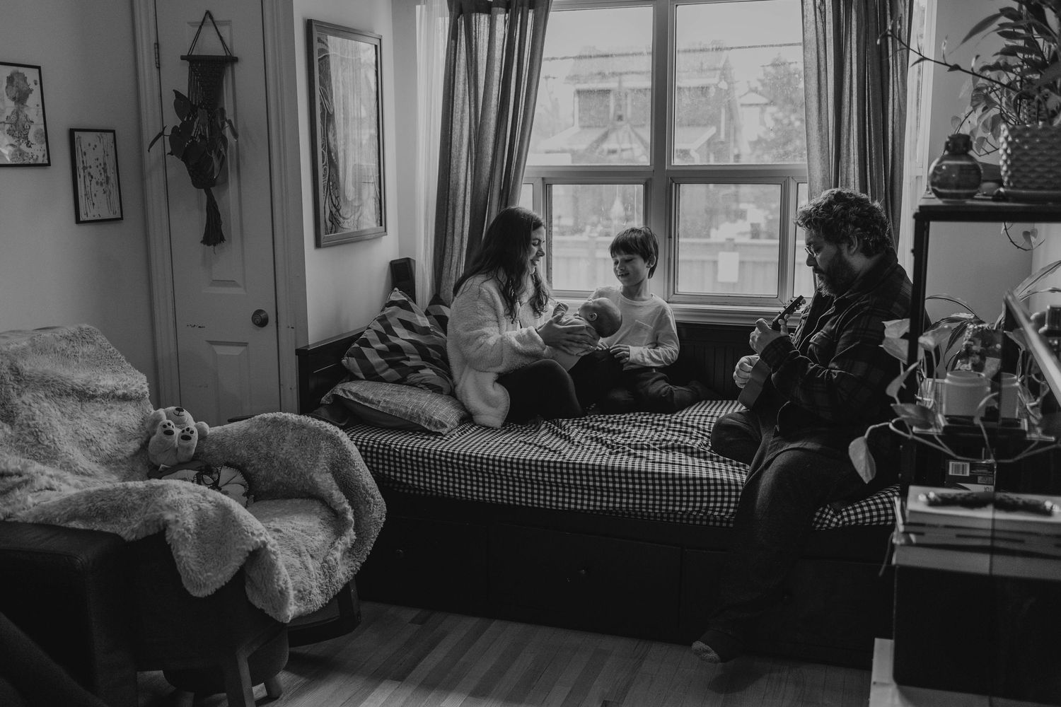 b&w image a mom showing the baby brother to the oldest close to the window in their sofa, while father is playing a guitar