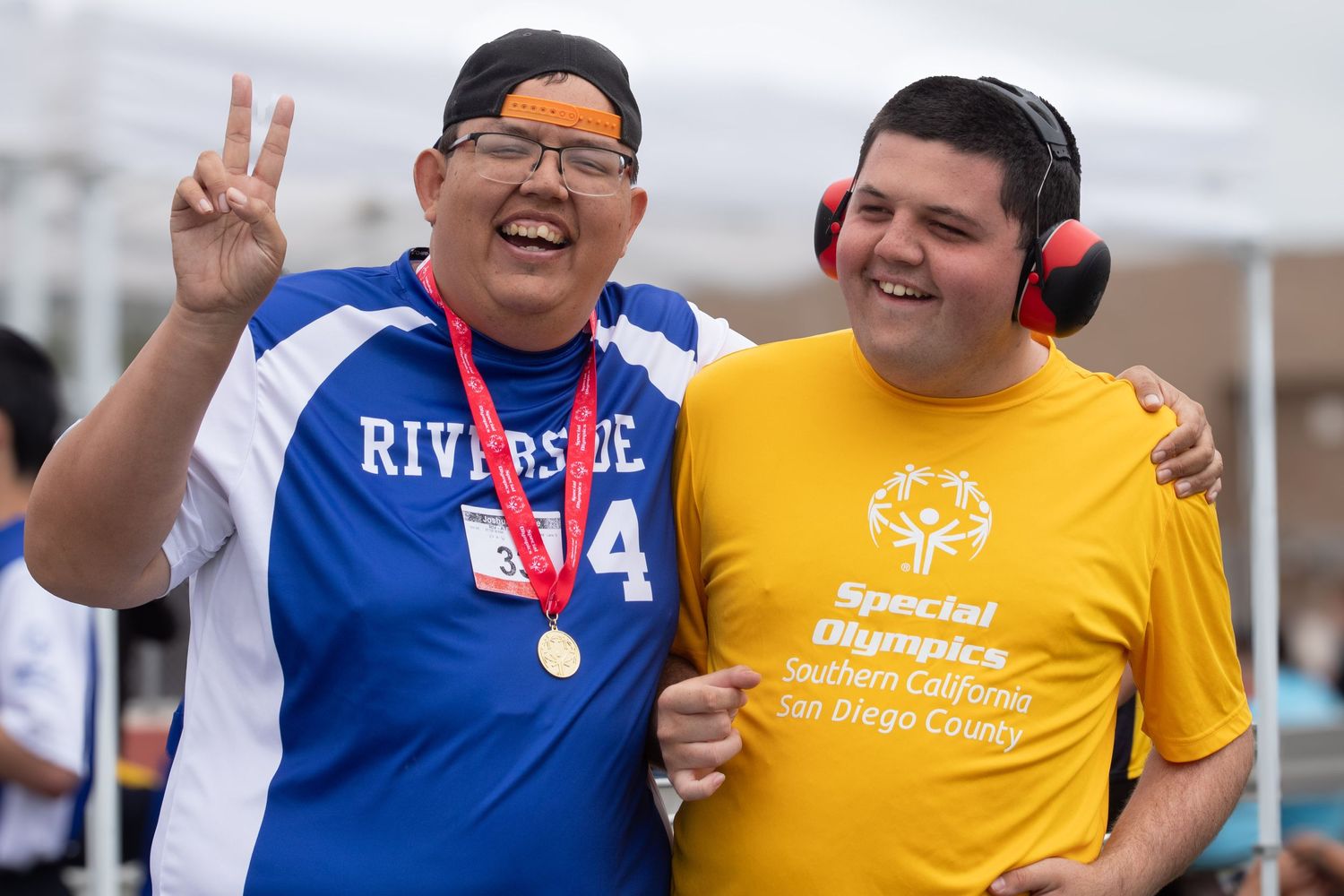 Two athletes at a Special Olympics event wearing blue and yellow shirts smile and make gestures while wearing medals.
