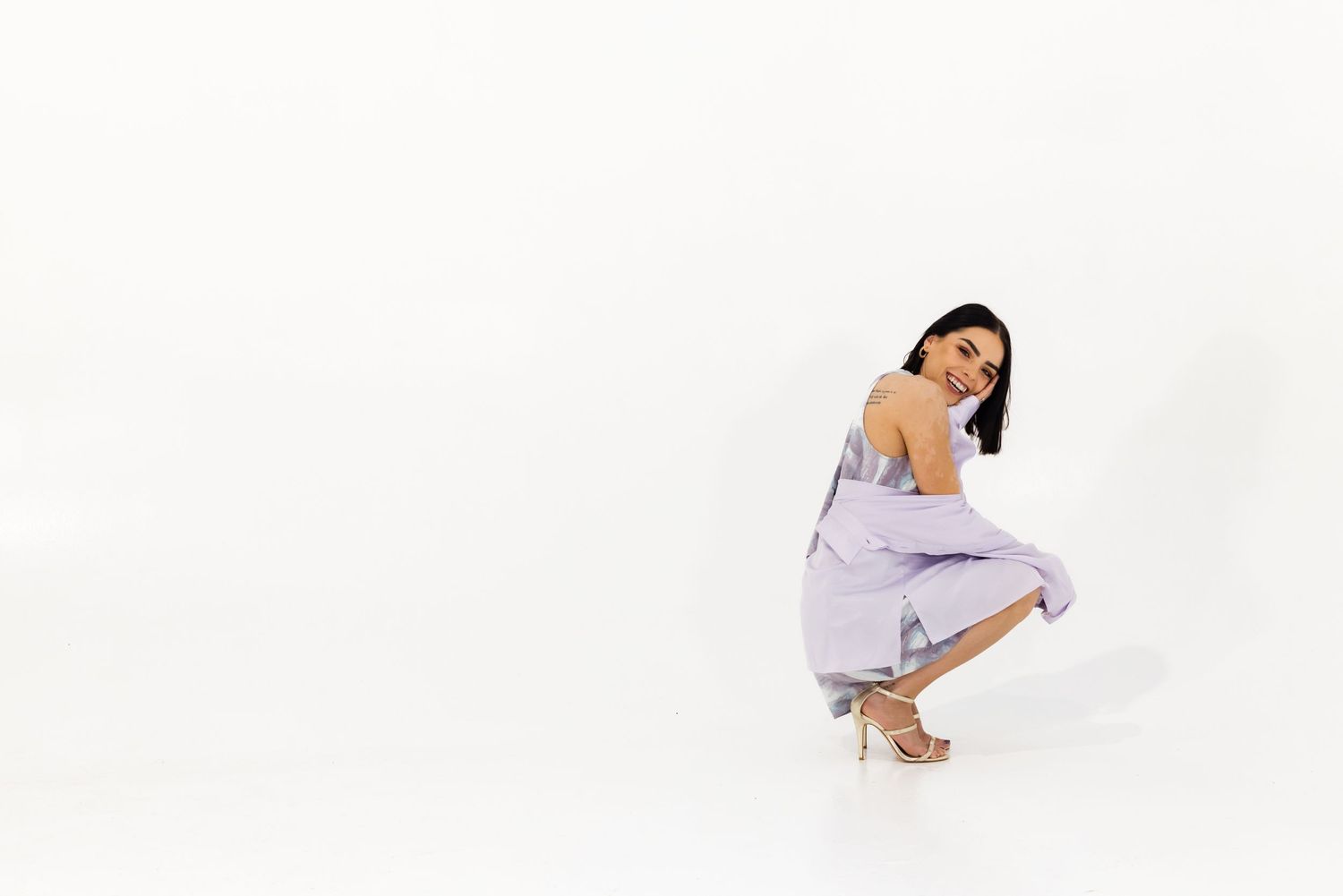 A person in a lavender dress and heels crouches elegantly against a white background in a fashion pose.