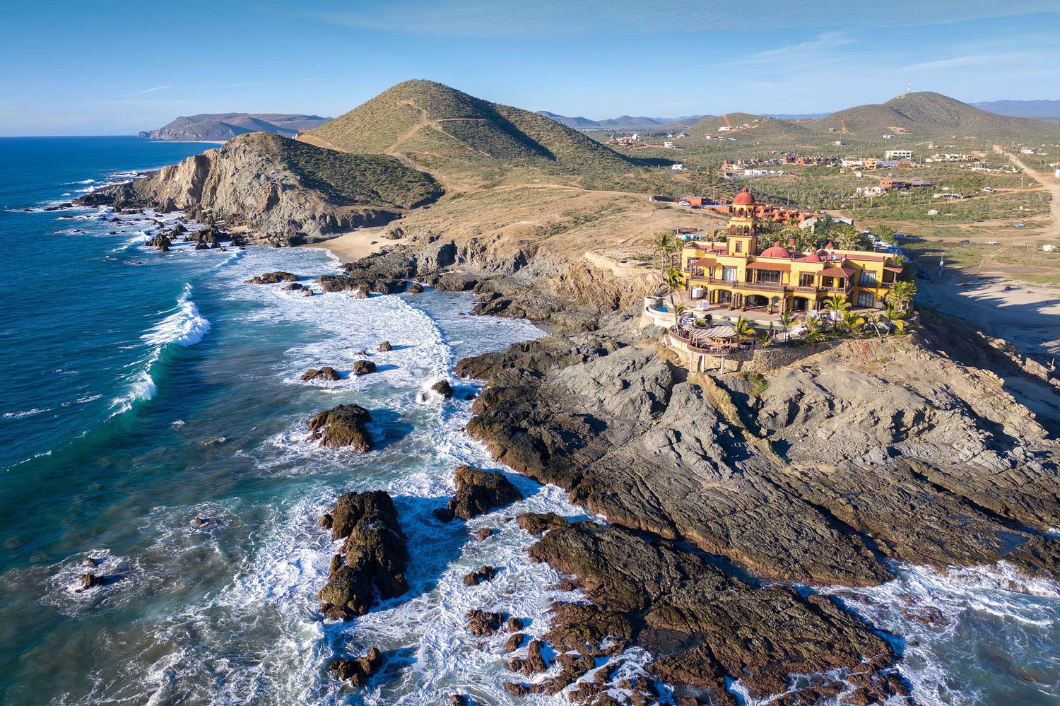 Dramatic aerial view of coastal landscape with rocky cliffs, crashing waves, and Spanish-style architecture. Hacienda Cerritos.