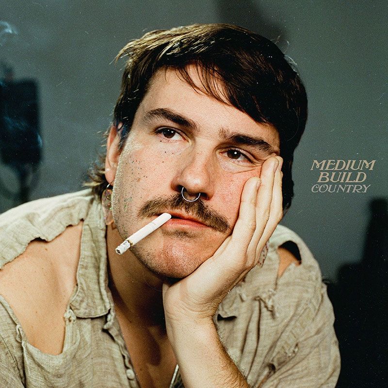A person with a prominent mustache rests their face on their hand while smoking a cigarette in a sepia-toned portrait.