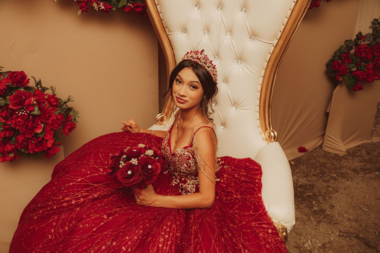 A quinceanera dress in deep burgundy red sparkles as someone poses on an elegant cream throne chair with roses.