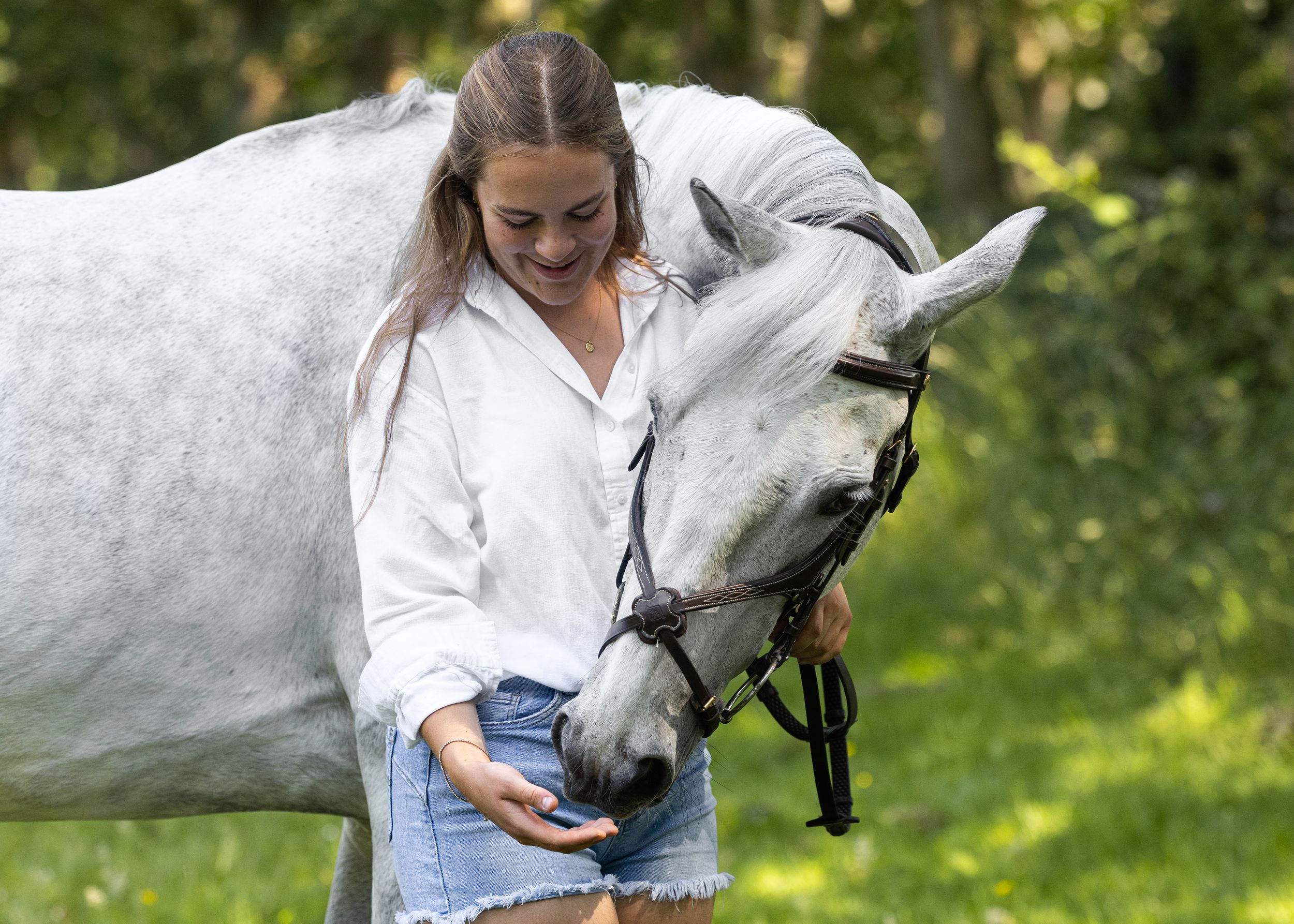 Young woman in white shirt feeding a gray horse outdoors in a lush green setting.