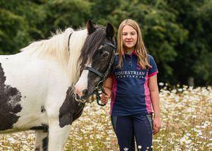 Young girl in equestrian shirt standing with black and white pinto horse in flower meadow.