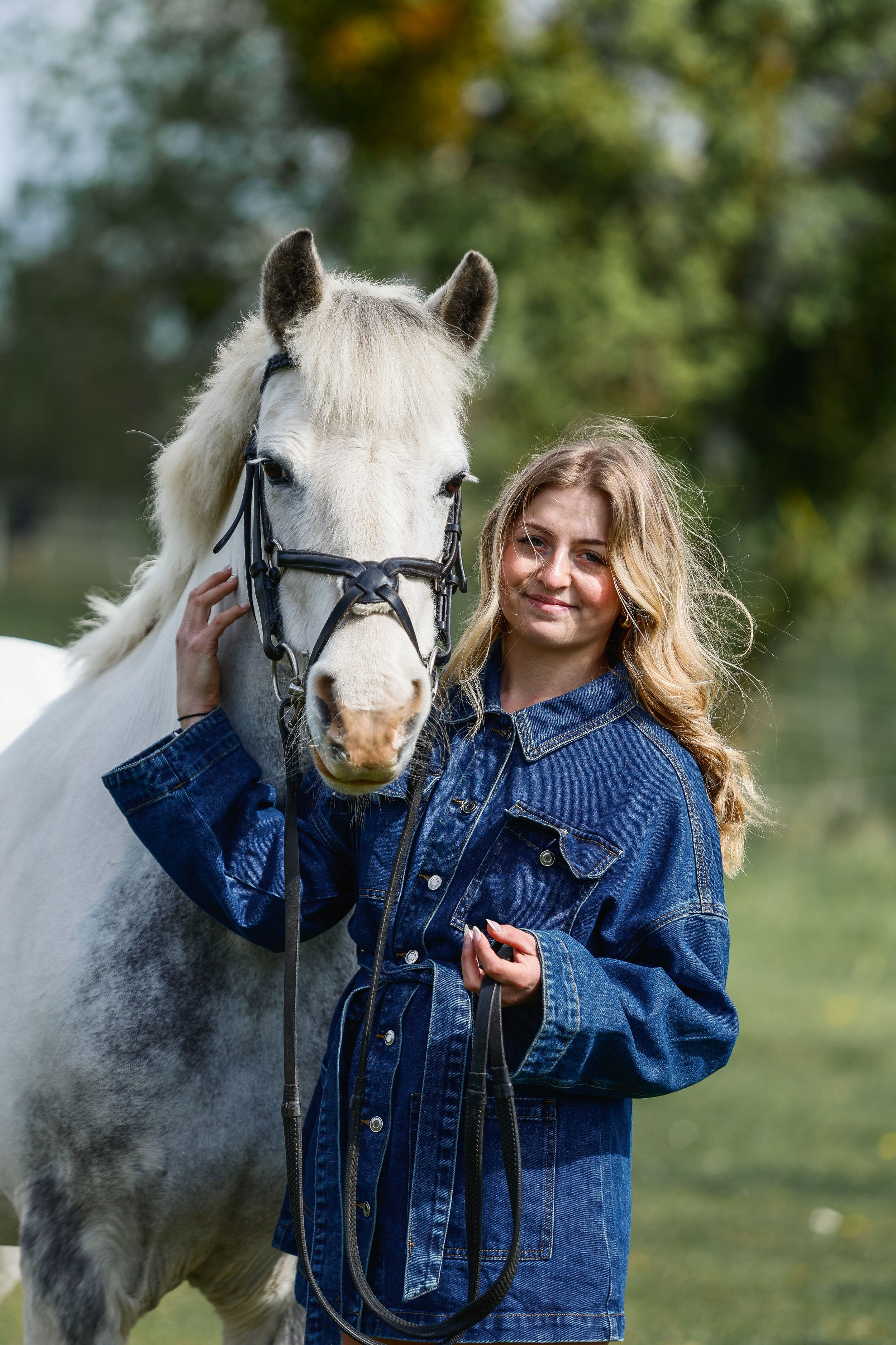 Young blonde woman in denim jacket smiling beside a white horse outdoors in a green field.