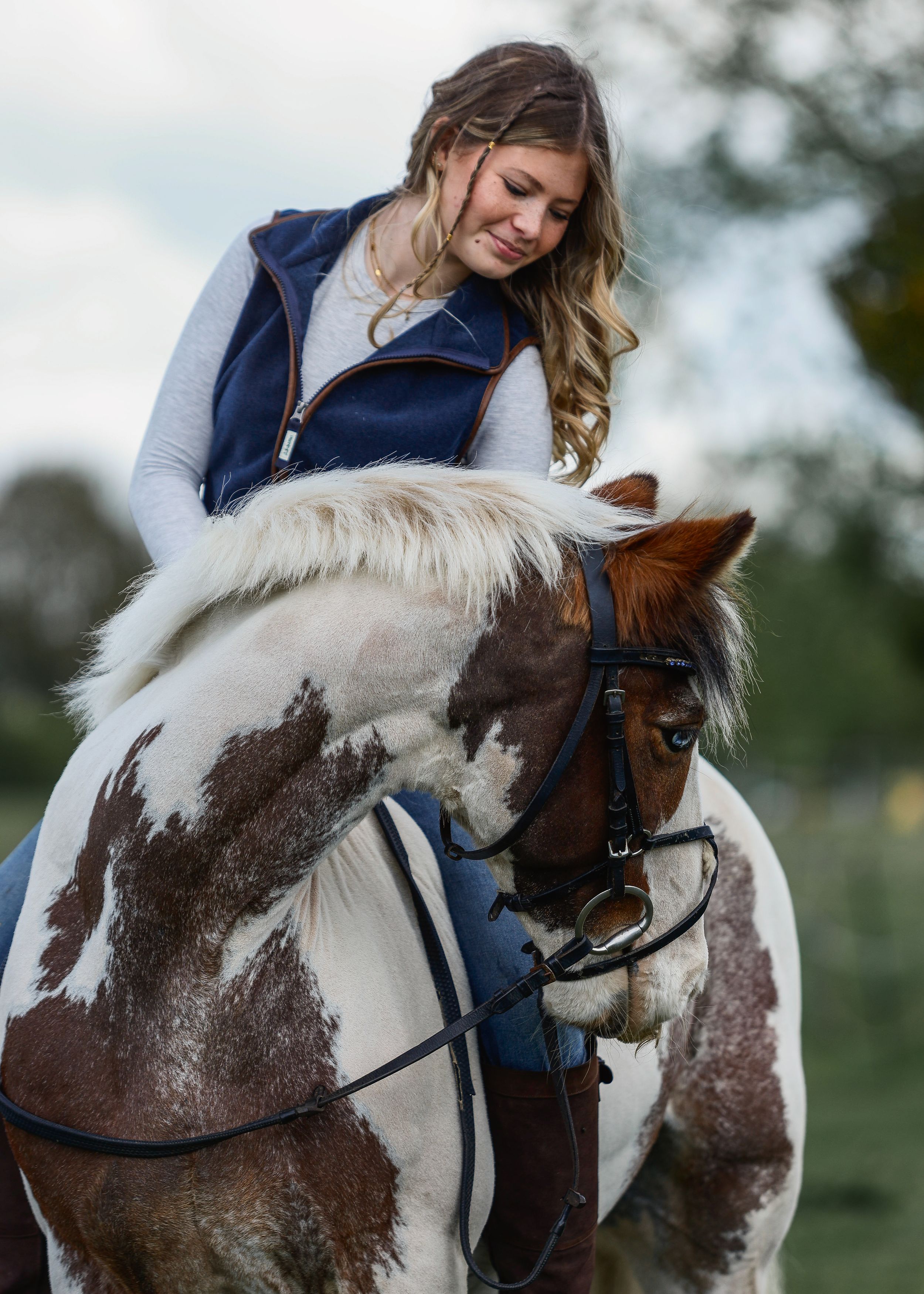 A young woman in a blue vest rides a brown and white paint horse outdoors, looking down affectionately.