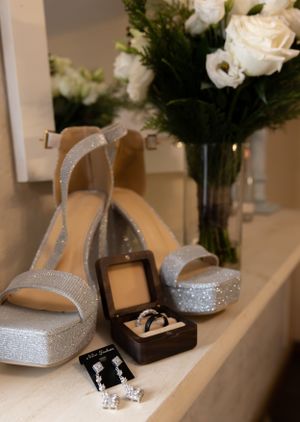 Sparkly silver bridal heels and wedding jewelry displayed next to a white rose bouquet in a vase.