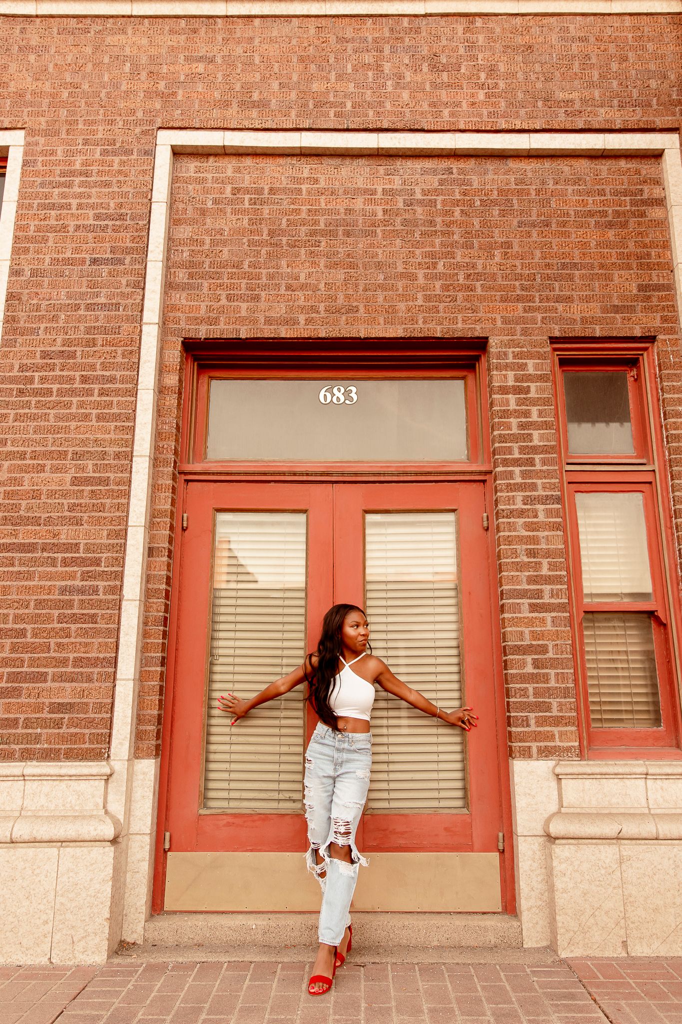 A person poses in casual summer attire against a red brick building with a red door marked 683.