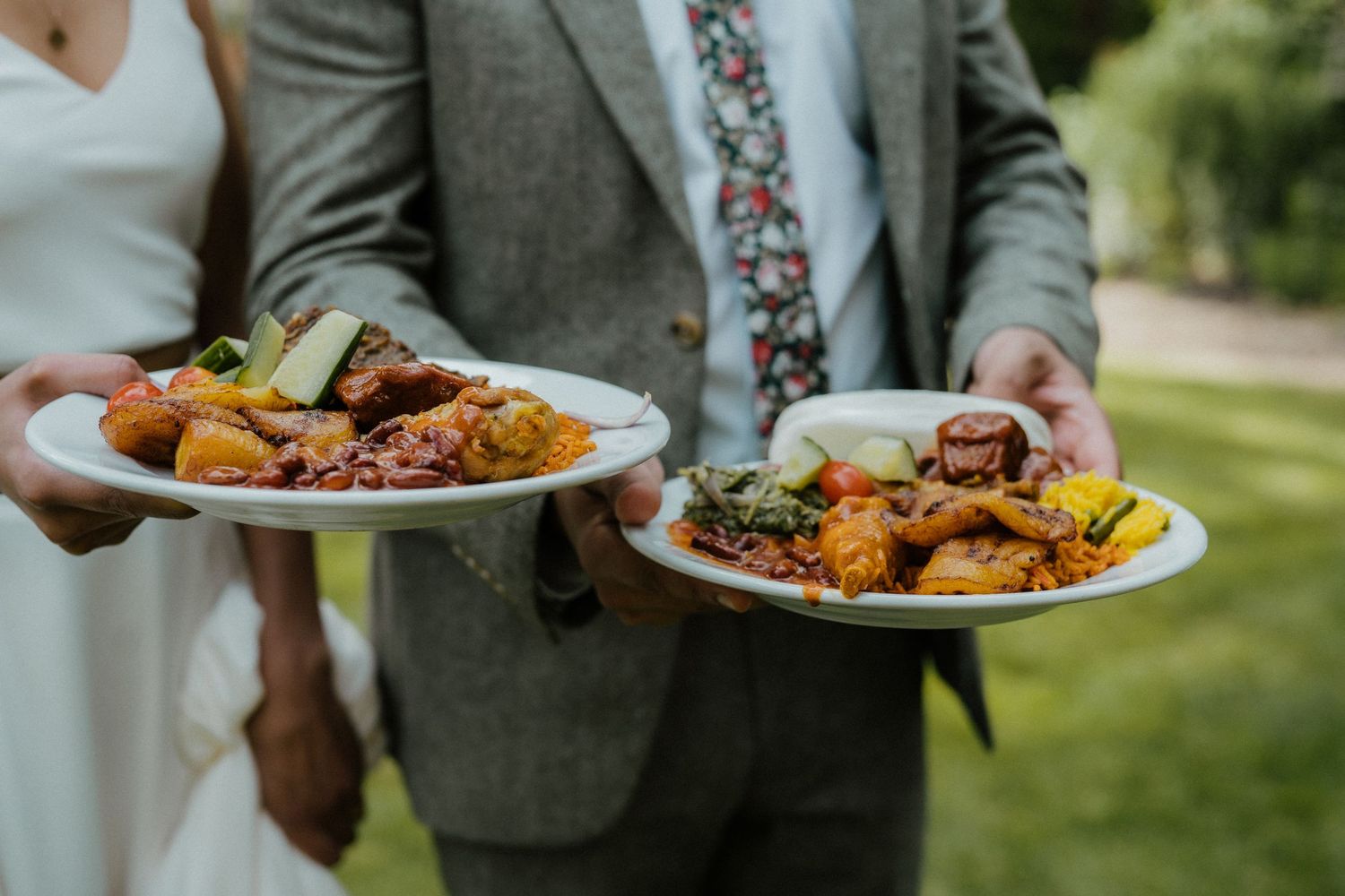 Two people in wedding attire hold plates of colorful plated meals with roasted meats and sides outdoors.